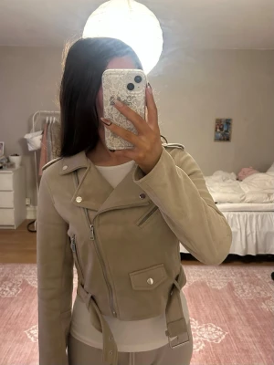 Beige mocka bikerjacka Stradivarius - Snygg beige bikerjacka i mjuk mockaimitation från Stradivarius. Jackan har flera silvriga dragkedjor, bred krage och bälte i midjan. Klassisk bikerstil med detaljer som tryckknappar och fickor. Perfekt för dig som vill ha en trendig och stilren look.