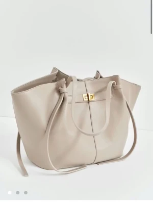 Beige bucket bag med gulddetaljer - Stilren beige bucket bag i slätt konstläder med långa axelband och snygg guldfärgad vridlåsdetalj framtill. Väskan har en rymlig form och minimalistisk design som passar till många olika looks. 