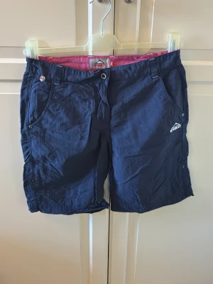 Marinblå shorts från McKinley - Snygga marinblå shorts från McKinley med klassisk passform och praktiska fickor. Shortsen har bälteshällor, knapp och dragkedja framtill samt en diskret logga broderad på ena benet. Storlek 36. Bra begagnad skick. Från McKinley