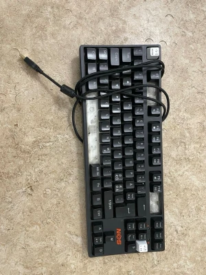 NOS mekaniskt tangentbord USB - Säljer ett svart mekaniskt tangentbord från NOS med USB-anslutning. Tangentbordet har nordisk layout med tydliga vita och gråa tangenter samt orange NOS-logga. Kompakt design och robust plastmaterial, perfekt för gaming eller skrivarbete.