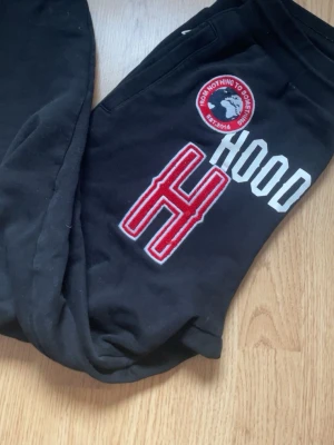 Svarta mjukisbyxor från Hoodrich - Svarta mjukisbyxor från Hoodrich med stor röd och vit H och texten HOOD på ena benet. Byxorna har resår i midjan och mudd vid benslut. Snygg detalj med rund patch och broderade bokstäver. Perfekta för en chill och street-inspirerad look. Är 171, å dem sitter bra i längden på mig😊