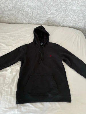  Polo Ralph Lauren Hoddie - Säljer en svart hoodie från Polo Ralph Lauren med klassisk huva och känguruficka. Den har det ikoniska röda Polo-märket broderat på bröstet. Tillverkad i mjukt material som känns skönt mot huden. Perfekt för en avslappnad och stilren look.