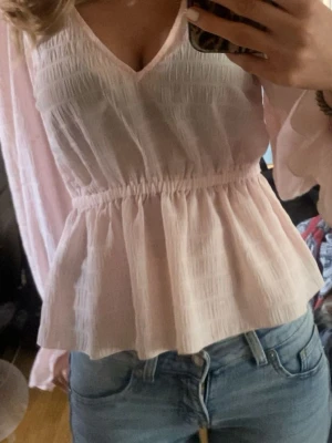 Ljusrosa blus med v-ringning - Söt ljusrosa blus med v-ringning och långa, lätt transparenta ärmar. Blusen har smockdetaljer över bröstet och midjan samt volang nedtill för en feminin siluett. Tillverkad i ett tunt, luftigt tyg som ger en romantisk känsla.