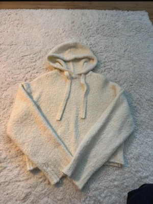 Krämvit fluffig hoodie h&m - Mysig krämvit hoodie med huva och dragsnören. Tillverkad i ett fluffigt, mjukt material som känns riktigt skönt mot huden. Hoodien har långa ärmar och en avslappnad passform, perfekt för chill dagar. 