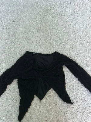 Svart omlottopp från SHEIN - Säljer en svart omlottopp från SHEIN med lång ärm och unik struktur i tyget. Toppen har en spetsig nederkant och är tillverkad i ett lätt, syntetiskt material. Perfekt för dig som vill ha något trendigt och edgy i garderoben!