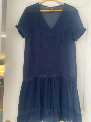 Marinblå kortklänning med spetsdetaljer - Söt marinblå kortklänning med v-ringning och spetsdetaljer framtill. Klänningen har korta ärmar och volang nedtill för en luftig känsla. Perfekt för varma dagar och enkel att styla med accessoarer.