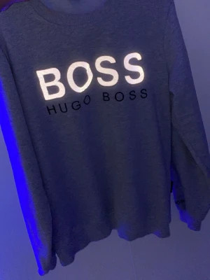 Grå sweatshirt från Hugo Boss - Säljer en grå sweatshirt från Hugo Boss med stort reflekterande BOSS-tryck och svart HUGO BOSS-text på bröstet. Tröjan har rund halsringning och långa ärmar. Perfekt för en avslappnad och stilren look.