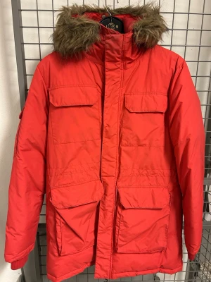 Röd parkas med fuskpälsluva - Röd parkas med fyra stora fickor framtill och huva med brun fuskpäls. Jackan har rak passform och dold dragkedja med vindslå. Perfekt för kalla dagar och har en klassisk streetstil.
