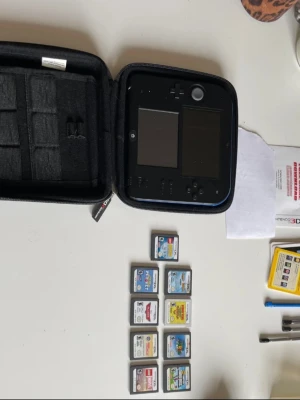 Nintendo 2DS med spel och fodral - Nintendo 2DS i svart med blå detaljer, två skärmar och klassiska knappar. Medföljer ett svart skyddsfodral och nio spelkassetter, bland annat Super Mario, LEGO Marvel och Planes. Perfekt för dig som gillar bärbart spelande och Nintendo-klassiker.