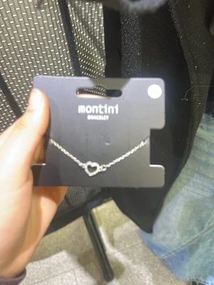 Silverfärgat armband med hjärta Montini - Snyggt silverfärgat armband från Montini med en tunn kedja och ett hjärta i mitten. Perfekt accessoar för att ge din outfit en söt touch. Justerbar längd med karbinhake. Passar till många olika stilar.