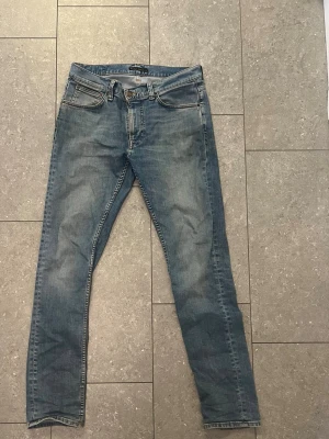 Blå lean dean jeans från Nudie Jeans - Säljer ett par klassiska blå jeans från Nudie Jeans. Jeansen har fem fickor, dragkedjegylf och subtila slitningar för en avslappnad look. Tillverkade i mjuk denim med Nudie-logga på bakfickorna. Perfekta till en streetwear-outfit. Modellen heter lean Dean och är storlek w32 L34. Hör av er vid frågor eller funderingar!!
