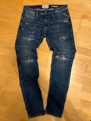 Replay anbas med slitningar - Säljer ett par blå jeans från Replay med snygga slitningar. Modellen har smal passform, Klassisk denimlook med feta slitningar på framsidan och bakfickor med broderade detaljer.