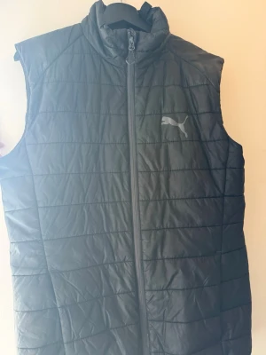 Svart väst från Puma - Svart quiltad dunväst från Puma med hög krage och dragkedja framtill. Västen har en stilren look med diskret Puma-logga på bröstet och nedtill på ryggen. 