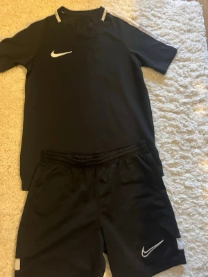 Svart Nike Dri-Fit träningsset - Säljer ett svart träningsset från Nike med Dri-Fit-teknologi. Setet består av en kortärmad tröja och matchande shorts, båda med vita Nike-loggor. Perfekt för fotboll eller gym. Materialet är lätt och andas bra.