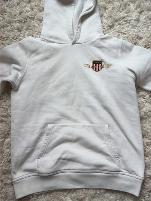 Vit hoodie från Gant med logga - Säljer en vit hoodie från Gant med broderad logga och sköld på bröstet. Hoodien har en klassisk känguruficka framtill och ribbade muddar vid ärmslut och nederkant. Tillverkad i mjukt bomullsmaterial och har en avslappnad passform med huva.