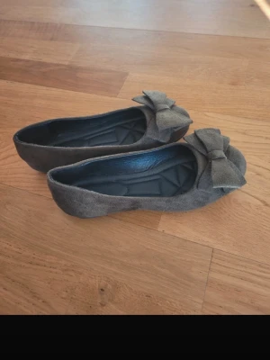 Gröna ballerinaskor med rosett - Säljer ett par gröna ballerinaskor i mockaimitation med stora dekorativa rosetter framtill. Skorna har en klassisk rundad tå och mjuk innersula för extra komfort. Perfekta för dig som gillar en söt och stilren look.