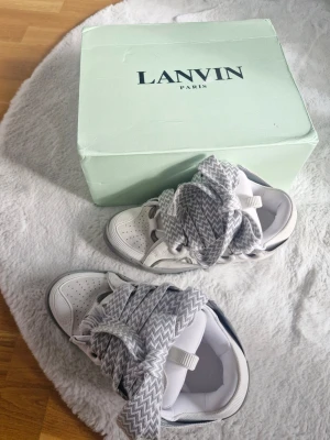 Lanvin sneakers med breda snören - Säljer ett par vita sneakers från Lanvin med breda, gråvita snören i sicksackmönster. Skorna har perforerade detaljer framtill och en klassisk sneakerform. Kommer med originalkartong i ljusgrönt. Perfekta för dig som gillar statement-detaljer.