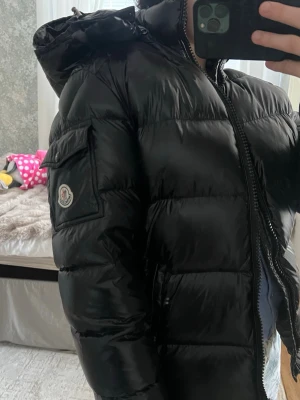 Svart dunjacka från Moncler - Säljer en svart, glansig dunjacka från Moncler med huva och dragkedja framtill. Jackan har ett Moncler-märke på ärmen och en ficka med lock på sidan. Perfekt för kalla vinterdagar och riktigt snygg att bära till jeans eller joggers. Använd ca 4 gånger