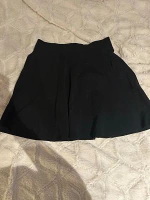 Svart kortkjol från H&M Divided - Enkel och stilren svart kortkjol från H&M Divided. Kjolen har en lätt utställd form och bred linning som ger en snygg siluett. Perfekt att matcha med en crop top eller stickad tröja för en trendig look.