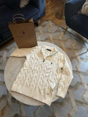 Vit kabelstickad tröja Polo Ralph Lauren - Kabelstickad vit tröja från Polo Ralph Lauren med halv dragkedja och ribbade muddar. Tröjan har det klassiska broderade logotypen på bröstet och en hög krage. Perfekt för en stilren och avslappnad look.