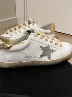 Golden Goose vita sneakers med stjärna - Snygga vita sneakers från Golden Goose med silverfärgad stjärna på sidan och guldfärgad häl. Beigea skosnören och GGDB/SSTAR-tryck på sidan. Klassisk låg modell i läder som passar perfekt till streetwear och casual outfits.