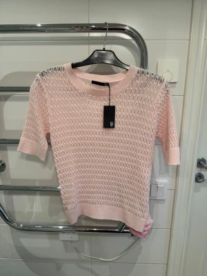 Ljusrosa stickad topp från 157 - Säljer en ljusrosa stickad topp från 157 med korta ärmar och rund hals. Tröjan har ett luftigt, hålmönstrat stickat material och ribbade kanter vid ärmslut och nederkant. Perfekt för lager på lager och en söt detalj till din outfit.
