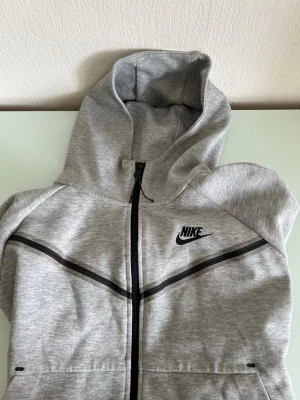 Grå zip hoodie från Nike - Snygg grå hoodie från Nike med dragkedja framtill och svart Nike-logga på bröstet. Hoodien har svarta detaljer längs bröstet och en stor huva. Tillverkad i mjukt sweatshirtmaterial, perfekt för en sportig och avslappnad stil.
