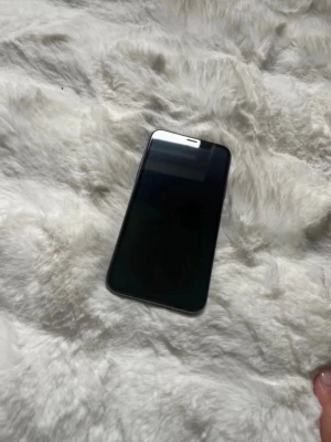 Svart smartphone med glasfront - Snygg svart IPhone XS med slimmad design och glasfront. Telefonen har rundade hörn och metallram, vilket ger en modern känsla. Perfekt för dig som vill ha en stilren mobil med stor skärm och smidigt format. Små skavanker runt sidorna och nån pytte liten repa på skärmen.