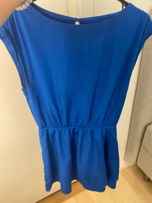 Blå enkel kortklänning - Säljer en stilren blå kortklänning med rund halsringning och resår i midjan. Klänningen har ett litet knappslå bak i nacken och är ärmlös, vilket ger en luftig och bekväm känsla. Perfekt för varma dagar och enkel att styla med accessoarer.