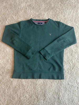 Mörkgrön sweatshirt från GANT - Mörkgrön sweatshirt från GANT med rund halsringning och diskret broderad logga på bröstet. Tröjan har långa ärmar och ribbade muddar vid ärmslut och nederkant. Perfekt för en avslappnad och stilren look.
