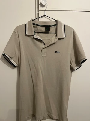 Beige pikétröja från BOSS oanvänd  - Klassisk beige pikétröja från BOSS med svart och vit rand på krage och ärmslut. Regular fit-modell med korta ärmar och diskret BOSS-logga broderad på bröstet. Perfekt till jeans eller chinos för en clean look.