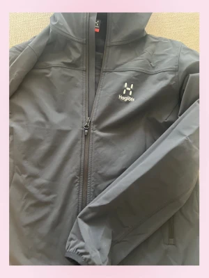 Svart vindjacka från Haglöfs - 💞Snygg svart vindjacka från Haglöfs med huva och hel dragkedja framtill. Jackan har två sidofickor med dragkedja och diskret Haglöfs-logga på bröstet. Tillverkad i lätt och slitstarkt material, perfekt för blåsiga dagar.💕