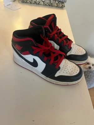 Nike Air Jordan 1 Mid svart/röd/vit - Säljer ett par Nike Air Jordan 1 Mid sneakers i svart, rött och vitt läder. Klassisk Swoosh på sidan, röda skosnören och perforerad tåbox för extra ventilation. Ikonisk retrostil som alltid är trendig.