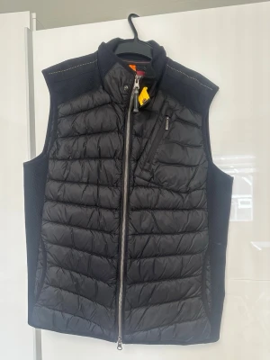 Svart dunväst från Parajumpers - Stilren svart dunväst från Parajumpers med quiltad design och dragkedja framtill. Västen har en bröstficka med dragkedja och detaljer i orange, samt ribbade partier på sidorna för extra komfort. Perfekt för lager-på-lager och streetwear.