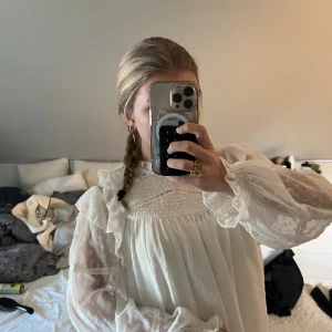 Vit romantisk blus med spetsdetaljer - Säljer en vit blus med transparenta, långärmade ärmar och fina spetsdetaljer. Blusen har volanger vid axlarna och hög krage med dekorativa sömmar. Perfekt för dig som gillar en romantisk och vintage-inspirerad stil.