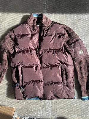 Brun dunjacka från Moncler - Snygg brun dunjacka från Moncler med stickade ärmar och krage. Jackan har två stora fickor framtill, dragkedja och ribbade detaljer i blått och vitt vid ärm- och nederkant. Perfekt för kalla vinterdagar och riktigt trendig look.