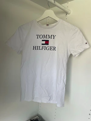 Tommy Hilfiger t-shirt - Klassisk vit t-shirt från Tommy Hilfiger med stort tryck och logga på bröstet. T-shirten har rund halsringning och korta ärmar. Tillverkad i mjuk bomull för en skön känsla och enkel stil.