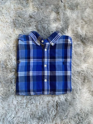 Blårutig skjorta från Ralph Lauren - Säljer en klassisk blårutig skjorta från Ralph Lauren med button-down krage och broderad logga på bröstet. Skjortan har vita knappar och är tillverkad i mjuk bomull. Perfekt för dig som gillar stilrena och tidlösa plagg.priset kan diskuteras vid snabb affär !!!!Den är i storlek L i barn!!!