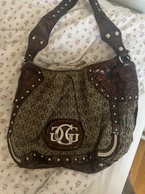 Brun och beige handväska från Guess - Vintage handväska från Guess i beige tyg med GG-logga över hela väskan och detaljer i mörkbrunt konstläder med croco-mönster. Väskan har bred axelrem med nitar och metallbeslag samt ett stort GG-emblem framtill. OBSERVERA ANVÄNT SKICK: materialet har börja lossna på remmarna. 