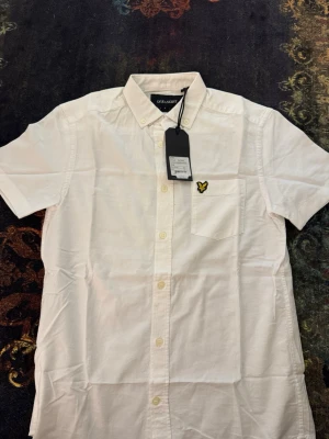 Vit kortärmad skjorta Lyle & Scott - Klassisk vit kortärmad skjorta från Lyle & Scott med knappstängning och bröstficka. Skjortan har en gul broderad logga på fickan och är tillverkad i lätt bomull. Perfekt till sommaren och enkel att matcha med olika outfits.