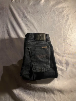 Mörkblå jeans från Nudie Jeans - Snygga mörkblå jeans från Nudie Jeans med klassisk femficksmodell och vågig söm på bakfickan. Jeansen har en svart läderpatch med logga i midjan och detaljer i gult. Perfekta för en avslappnad och trendig stil. W29/L30