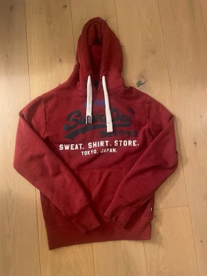 Röd hoodie från Superdry med tryck - Snygg röd hoodie från Superdry med stort tryck på bröstet och texten 'Sweat. Shirt. Store. Tokyo, Japan.' Mjuk insida, med huva, med vita snören och känguruficka framtill. Perfekt för dig som gillar streetwear och vill ha en bekväm och stilren look. Hör av dig om du vill ha fler bilder eller undrar något annat mvh Levi.