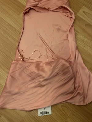 Ljusrosa satin klänning från NA-KD - Säljer en ljusrosa långklänning från NA-KD i mjukt satinmaterial. Klänningen har en öppen rygg med snörning och smala axelband. Perfekt för dig som vill ha en elegant och trendig look med en feminin touch.