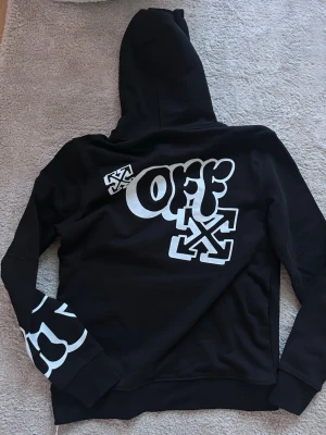 Svart Off-White hoodie med tryck - Svart hoodie från Off-White med stora vita tryck på ryggen, ärmen och fickan. Klassisk huva med snörning och Off-White-logga på bröstet. Perfekt för dig som gillar streetwear och vill sticka ut med en unik design.
