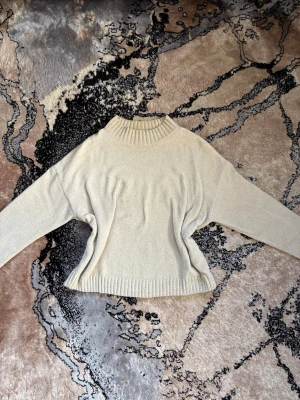 Beige stickad tröja med hög krage - En stilren beige stickad tröja med hög ribbad krage och ribbade muddar vid ärmslut och nederkant. Tröjan har en avslappnad passform och är perfekt för kyliga dagar. Materialet känns mjukt och bekvämt mot huden. Anledningen till att jag säljer tröjan är på grund av att jag börjar tröttna på den. 