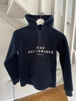 Mörkblå teddyhoodie Peak Performance - Mysig mörkblå teddyhoodie från Peak Performance med stor huva och halv dragkedja framtill. Hoodien har en stor magficka och texten 'PEAK PERFORMANCE ORIGINAL' broderad på bröstet. Perfekt för kalla dagar och chill outfits.