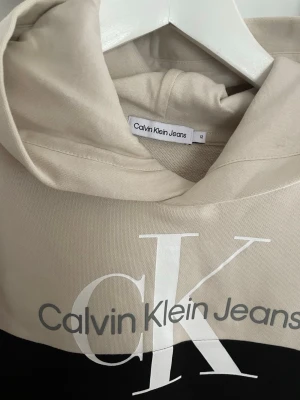 Beige och svart hoodie Calvin Klein - Snygg hoodie från Calvin Klein Jeans i beige och svart med stor CK-logga och text på bröstet. Hoodien har huva och långa ärmar. Tillverkad i mjukt bomullsmaterial som känns skönt mot huden. Perfekt för en avslappnad och trendig look.