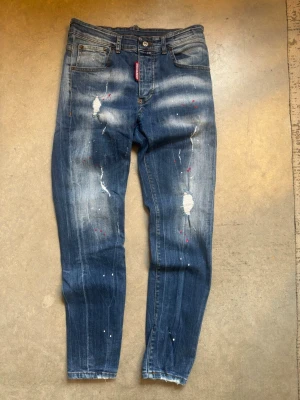 Dsquared2 jeans - Säljer ett par blå jeans från Dsquared2 med slitna detaljer, färgstänk och en cool tvätt. Jeansen har smal passform, klassisk femficksdesign och en röd Dsquared2-tag vid gylfen. Perfekta för dig som gillar en edgy streetstil.
