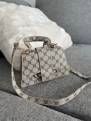 Mönstrad handväska - Snygg beige handväska med klassiskt GG-monogram i mörkbrunt. Väskan har både handtag och axelrem i samma mönstrade material samt guldfärgade metalldetaljer. Stängs med magnetlås och har svart innerfoder. Perfekt för dig som gillar lyxig och trendig design.