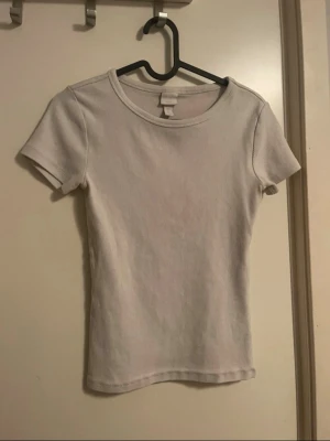 Vit ribbad t-shirt från H&M - En enkel och stilren vit t-shirt från H&M med ribbad struktur. Modellen är kortärmad och har en klassisk rund halsringning. Perfekt basplagg som passar till det mesta och är tillverkad i mjuk bomull för skön komfort.